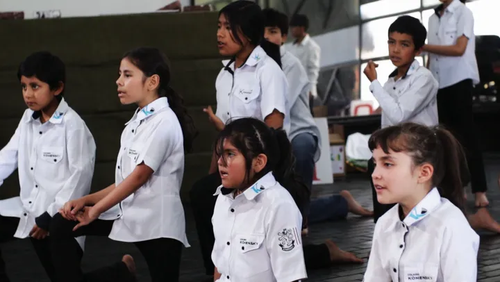 Alumnos ensayan una escena de rodillas, cantando, antes de subir al escenario